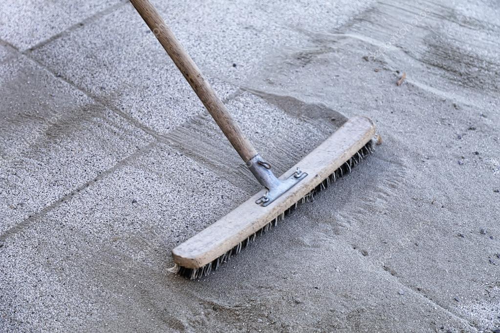 Wooden-handled push broom sweeping fine gravel/sand on a concrete surface; bristles flat against the ground.