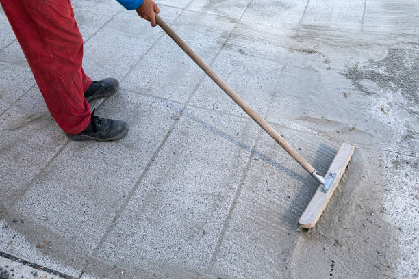 Person wearing red pants and a blue sleeve uses a long-handled wooden screed to level wet concrete on a paved surface.