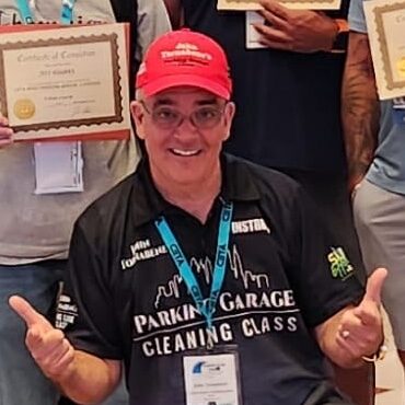Smiling man in a red cap and black polo reading 'Parking Garage Cleaning Class,' giving thumbs up at a certificate event.