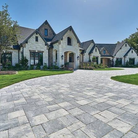 Stone-and-brick mansion with arched entry, expansive curved stone driveway, and manicured lawn under a clear blue sky.