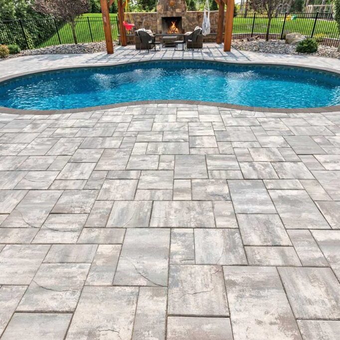 Kidney-shaped blue pool surrounded by a light gray stone patio, with a fire-pit seating area under a wooden pergola in a fenced backyard.